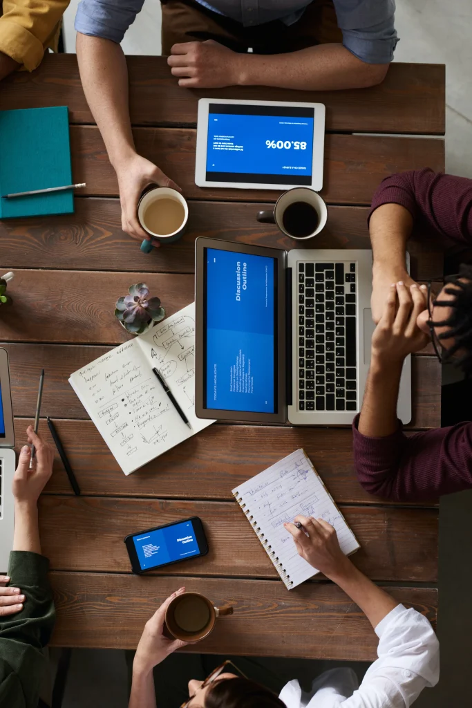 Blick von oben auf einen Besprechungstisch mit Laptops, Kaffeebechern und Notizbüchern, die in geordneter Weise angeordnet sind, während die Teilnehmer an kollaborativen Gesprächen teilnehmen